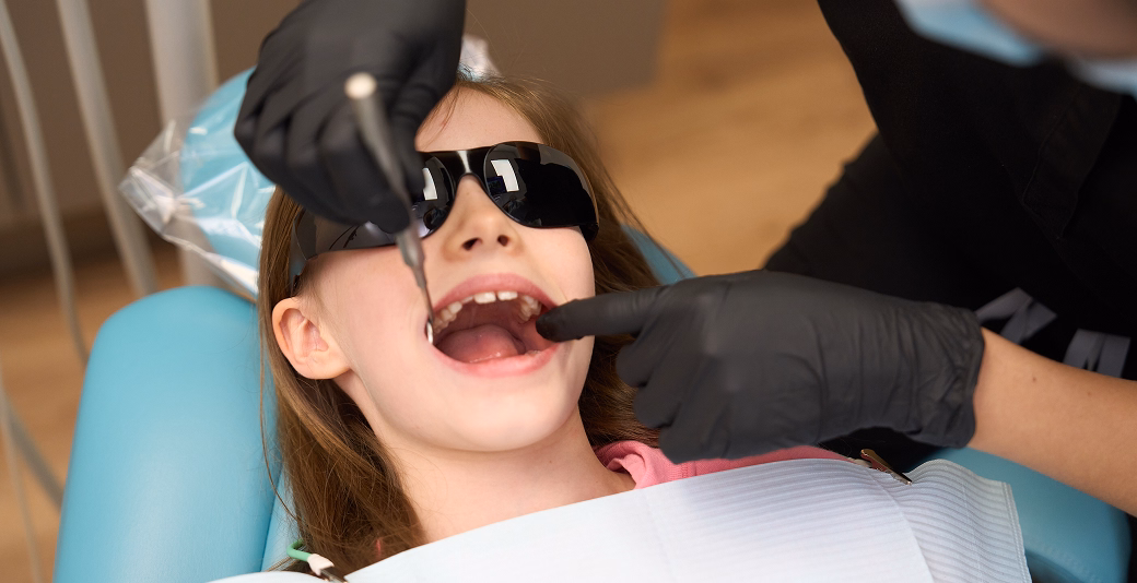Child receiving routine dental check-up, wearing protective sunglasses and bib, with dentist using tools in modern clinic setting.
