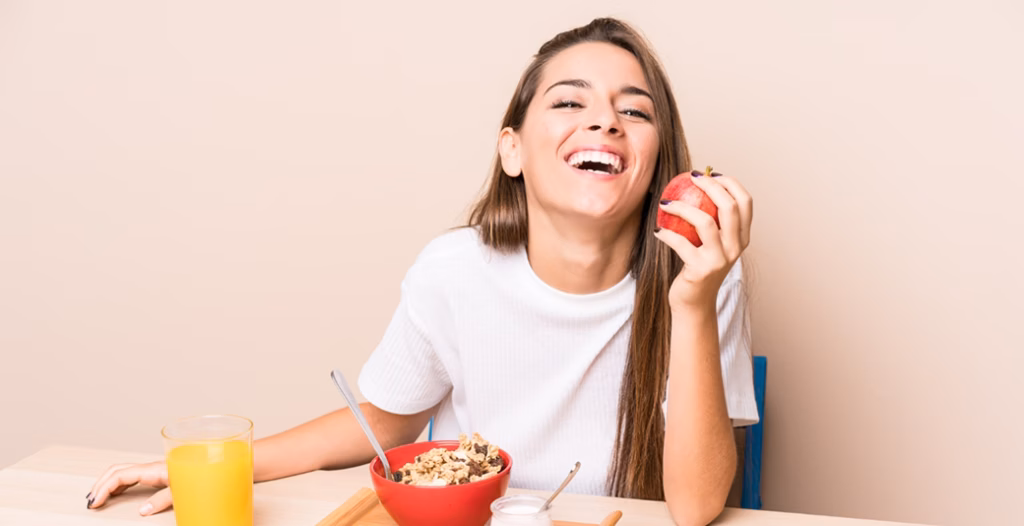 Smiling person holding a red apple at a breakfast table with granola, orange juice, yogurt, and a spoon in a red bowl.