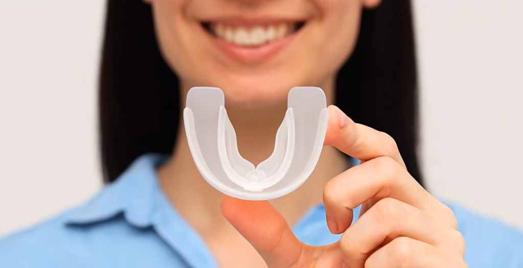 Smiling person holding a clear dental mouthguard in front of their face, wearing a light blue shirt.