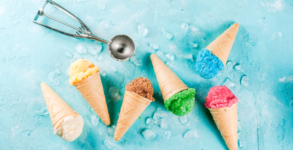 Six colorful ice cream cones arranged on blue surface with ice cubes and a metal scoop.