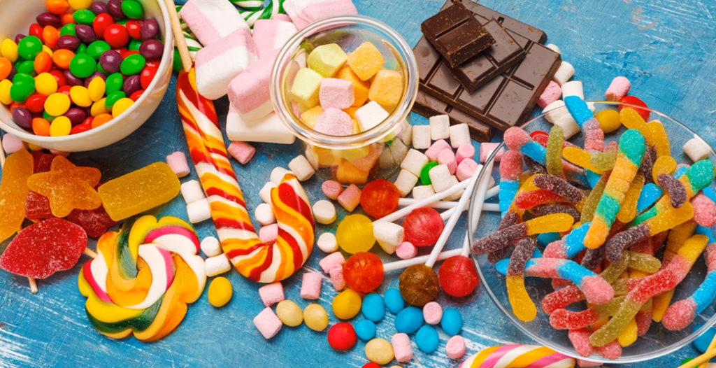 Colorful candies on blue wooden surface with gummy worms, lollipops, candy canes, marshmallows, and chocolates.