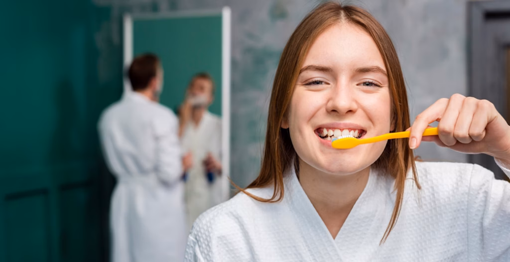 Person in white bathrobe brushing teeth with yellow toothbrush, another grooming in mirror in bathroom.