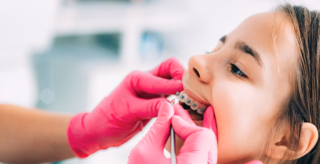 Orthodontist in pink gloves adjusting metal braces on young patient&rsquo;s teeth during procedure.