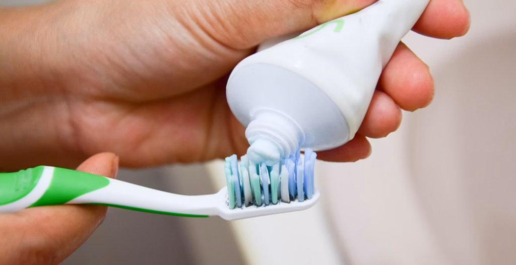 Light blue toothpaste being squeezed onto toothbrush with green-white handle and multicolored bristles.