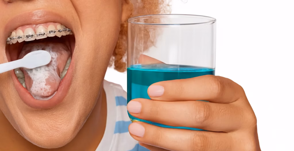 Person with braces brushing tongue while holding blue mouthwash, emphasizing complete oral hygiene routine.