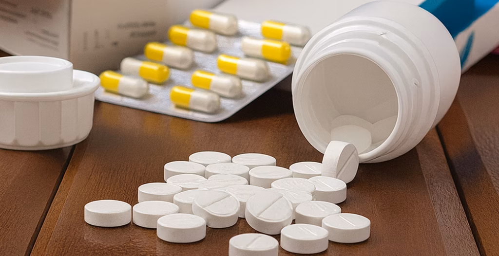 White tablets spilled from a pill bottle with blister packs and medicine boxes on a wooden surface.