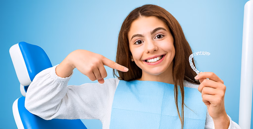 Teen in dental chair smiling, holding clear aligner, pointing to teeth, wearing bib in clinic setting.