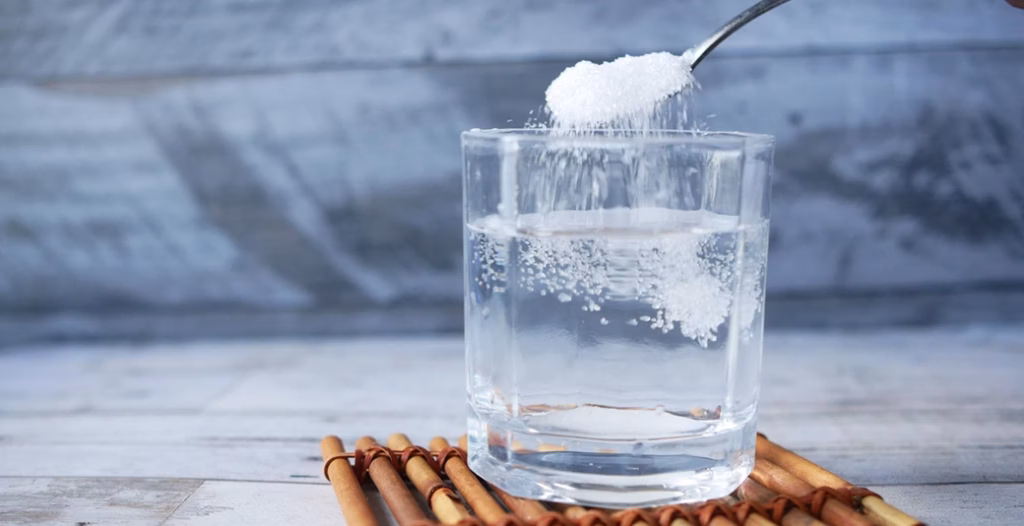 Clear glass of water on a bamboo mat with white granules falling from a spoon, dissolving into the warm water.