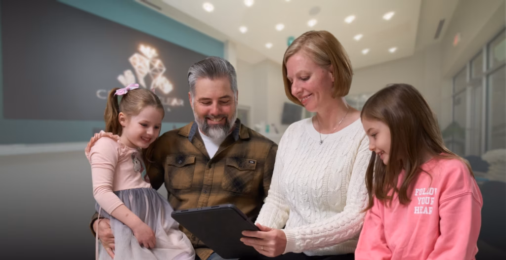 A family of four smiling together at Crystal Clear Dental, engaged with a tablet in a bright modern clinic.