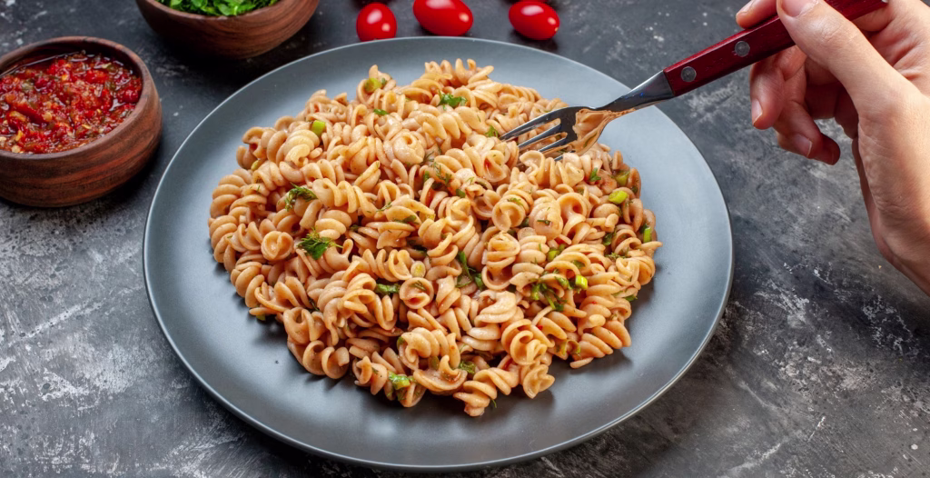 Plate of rotini pasta with herbs and vegetables, surrounded by sauce bowls and cherry tomatoes on rustic table.