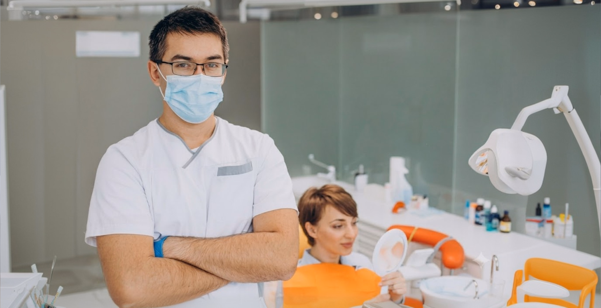 A male dentist with glasses and a mask stands with arms crossed in a clinic. A female patient with an orange bib holds a hand mirror in the background.