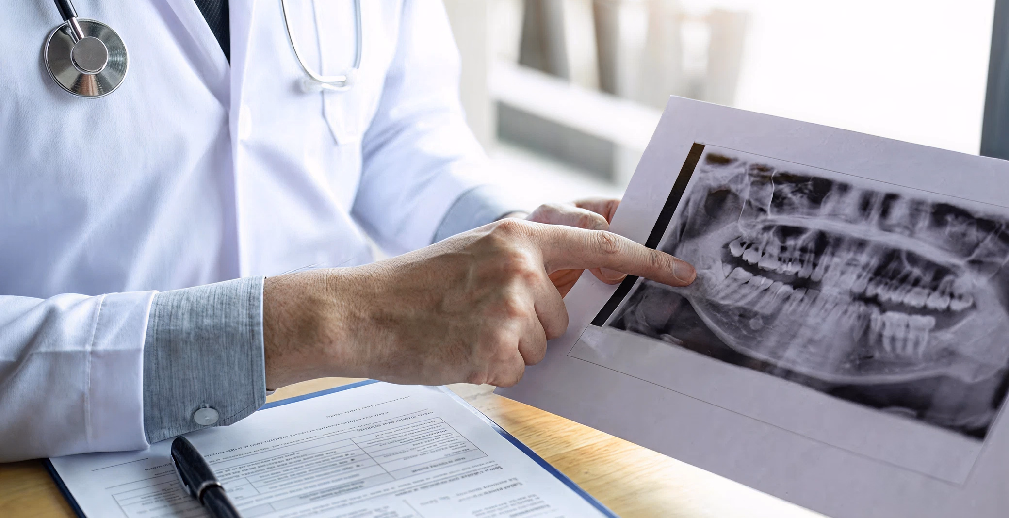 A doctor in a white coat points to a dental X-ray showing teeth and jaw, with a medical form on the desk.