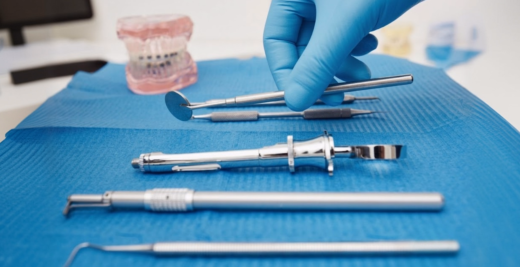 A gloved hand holds a dental mirror and explorer over other instruments and a model of teeth.