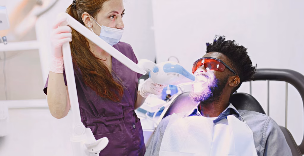 A dental assistant using a UV whitening light on a male patient wearing protective glasses.