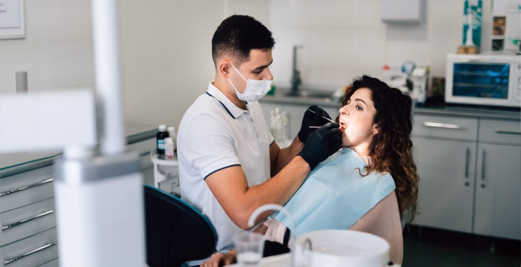 A dentist in a face mask and black gloves examines a curly-haired woman's teeth in a dental clinic room.