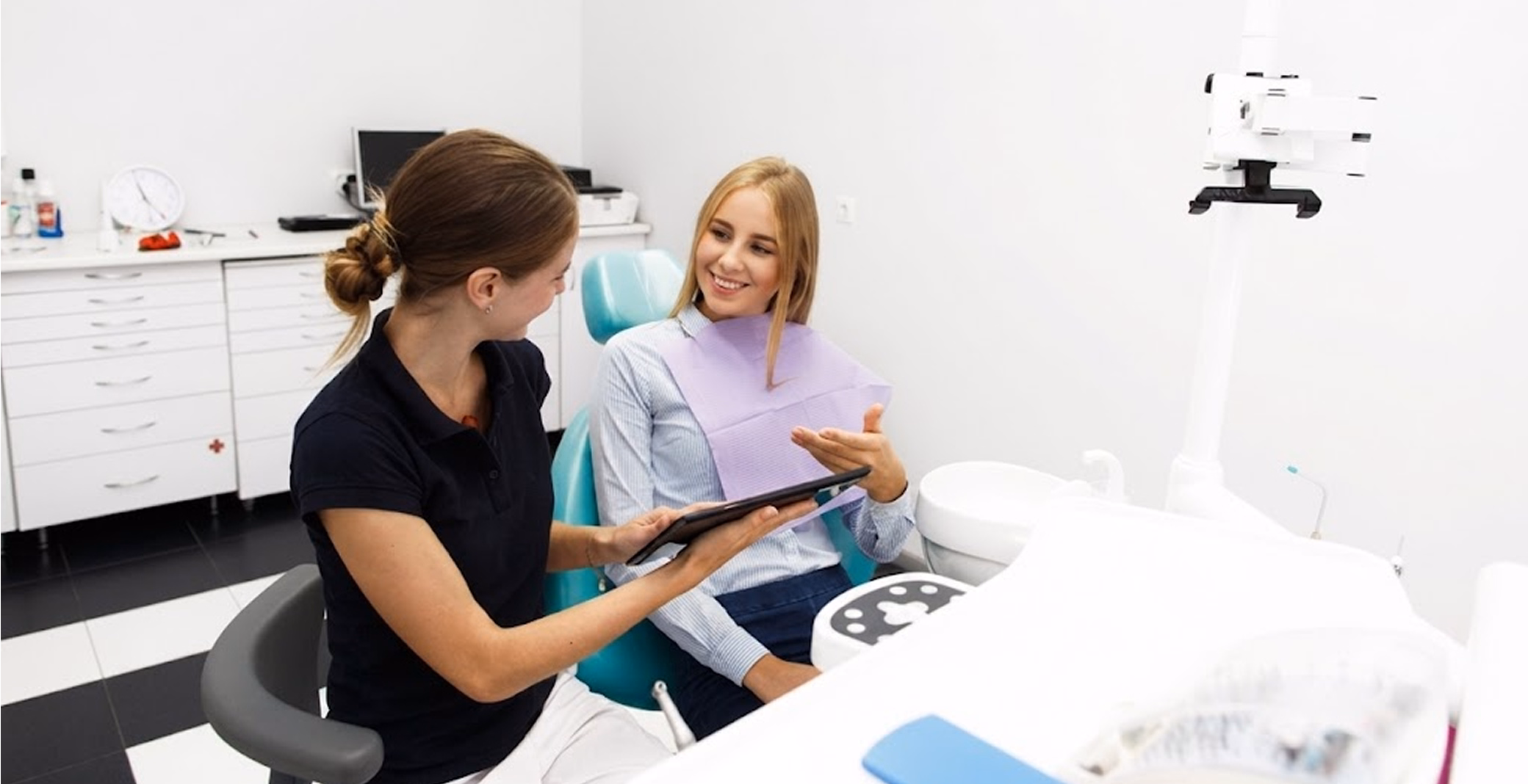 A woman in a blue shirt and dental bib looks at a tablet held by a dental professional, smiling as they sit together.