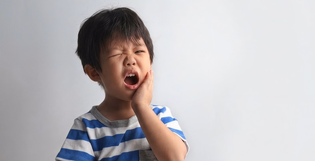 Young boy wincing in pain holding his cheek with an open mouth, wearing a blue and white striped shirt against light backdrop.