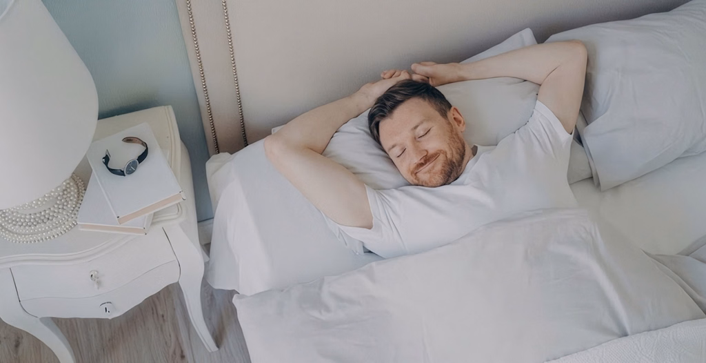 An overhead view of a smiling man with a beard sleeping peacefully on his back with his arms raised.