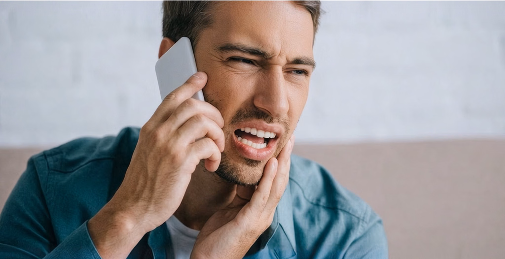 Man grimacing in pain while talking on a phone and holding his aching jaw.
